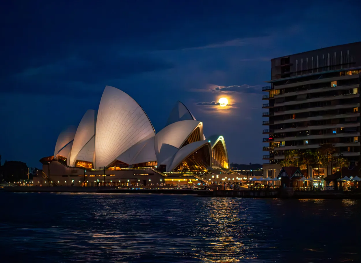Full moon rising behind Sydney Opera House at night, illuminated sails glowing against deep blue sky