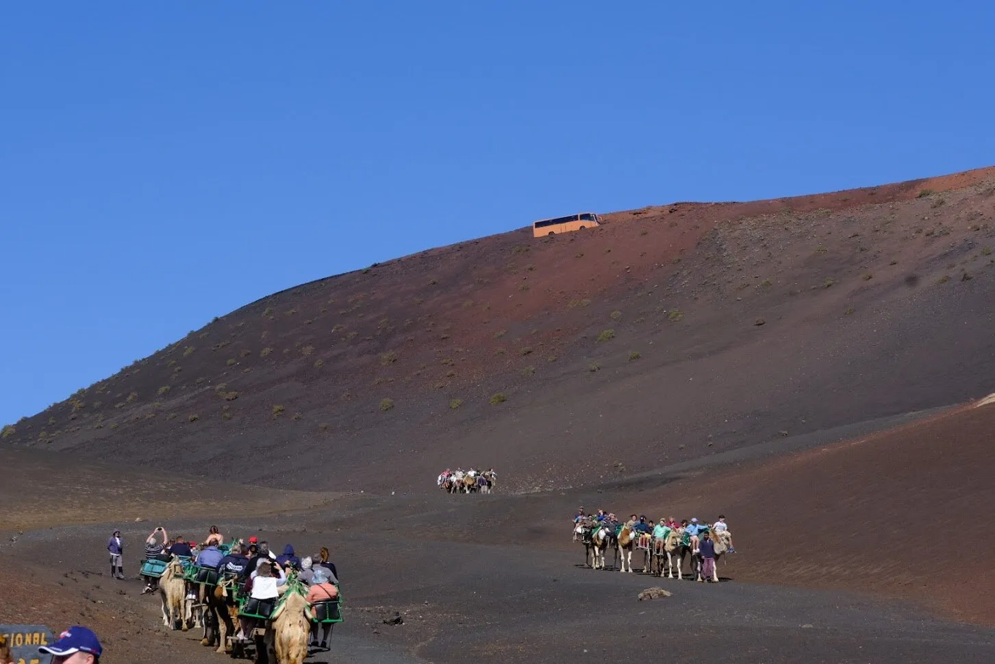 The yellow bus above the mountains and tourists on camel rides at Timanfaya National Park