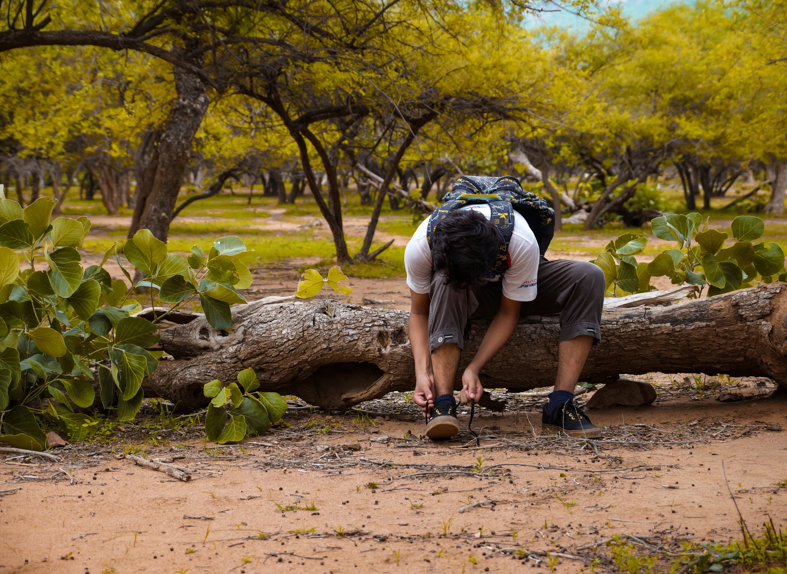 A boy sitting on a tree trunk tying his shoelaces